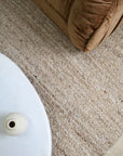 A close-up, high-angle view of a textured beige area rug in a living room setting, with the corner of a brown velvet sofa and a white side table visible.
