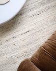 A close-up of a textured cream and beige area rug with subtle horizontal woven stripes, styled in a modern interior with a brown velvet chair and a white table edge visible.