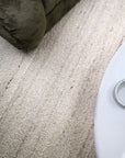 A close-up view of a textured, loop-pile area rug in a warm off-white or cream color, shown in a room setting with parts of a dark olive green upholstered piece of furniture and a white table visible.