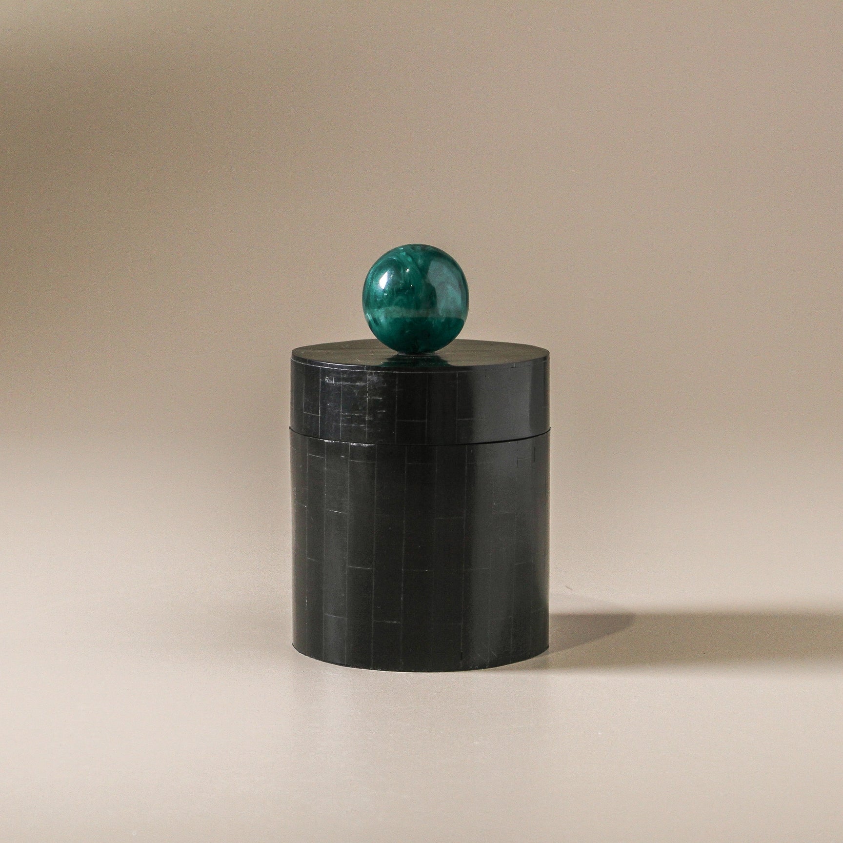 Black cylindrical container with a green marble ball on top against , perfect for storing trinkets.