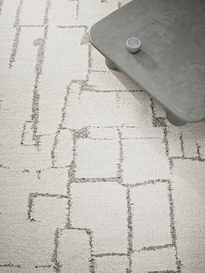A close-up of a light beige area rug with a distressed, geometric block pattern in dark gray. A gray concrete-style coffee table is partially visible in the top right corner.
