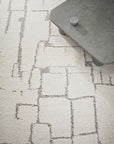 A close-up of a light beige area rug with a distressed, geometric block pattern in dark gray. A gray concrete-style coffee table is partially visible in the top right corner.