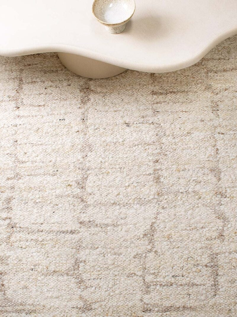 A close-up of a textured area rug in cream and beige with a subtle geometric pattern, styled under a modern white sculptural side table.