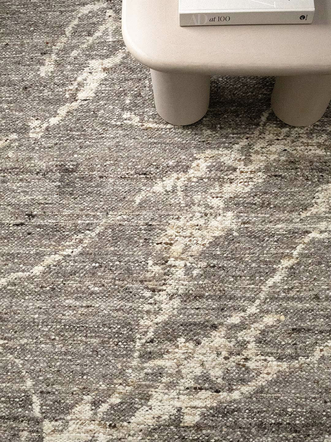 A close-up of a textured area rug with a distressed abstract pattern in gray and cream, with the lower portion of a light-colored stool visible at the top edge.