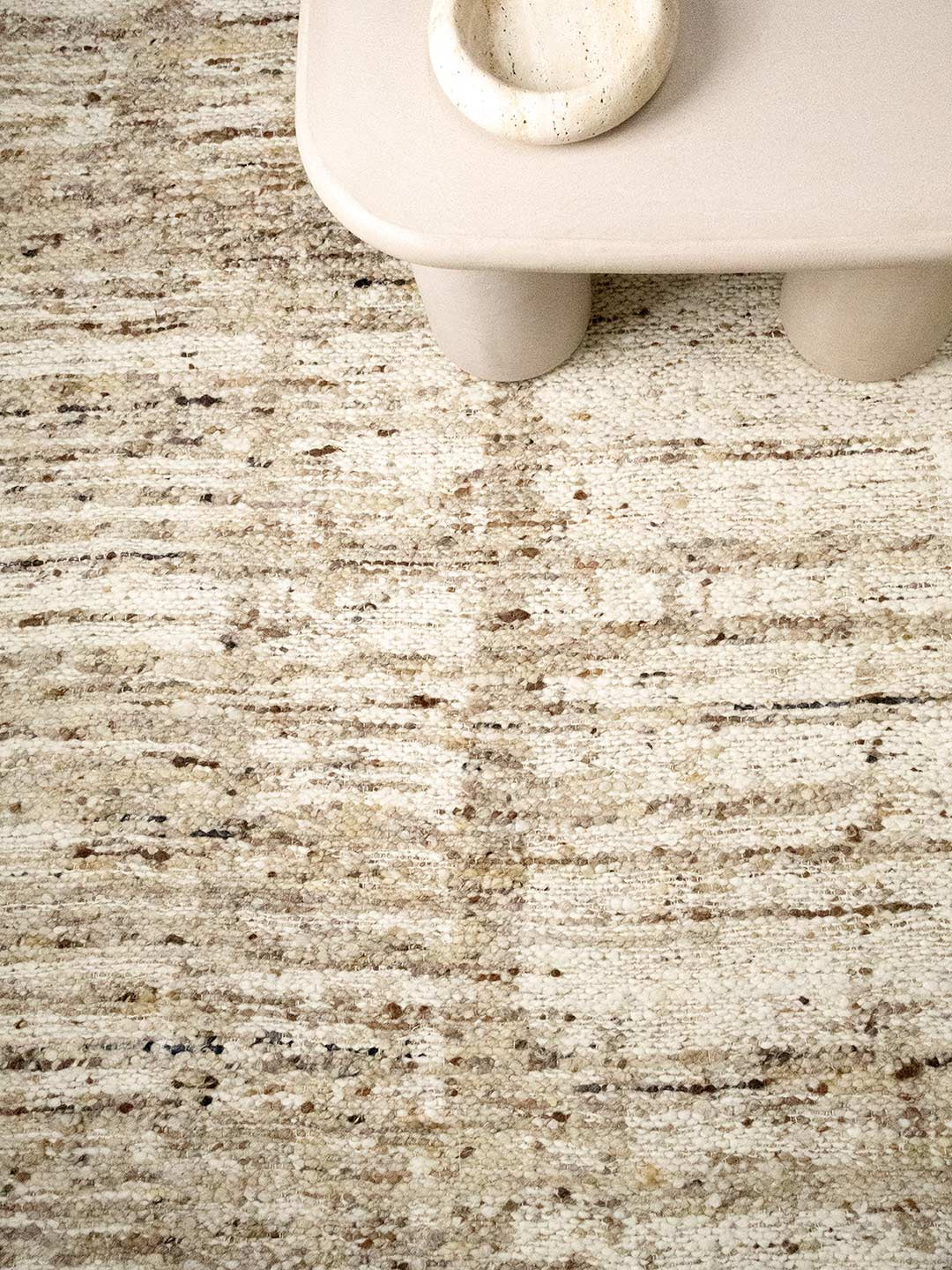 A close-up of a textured area rug in neutral beige, cream, and brown tones, with the lower edge of a light-colored wooden table visible at the top.