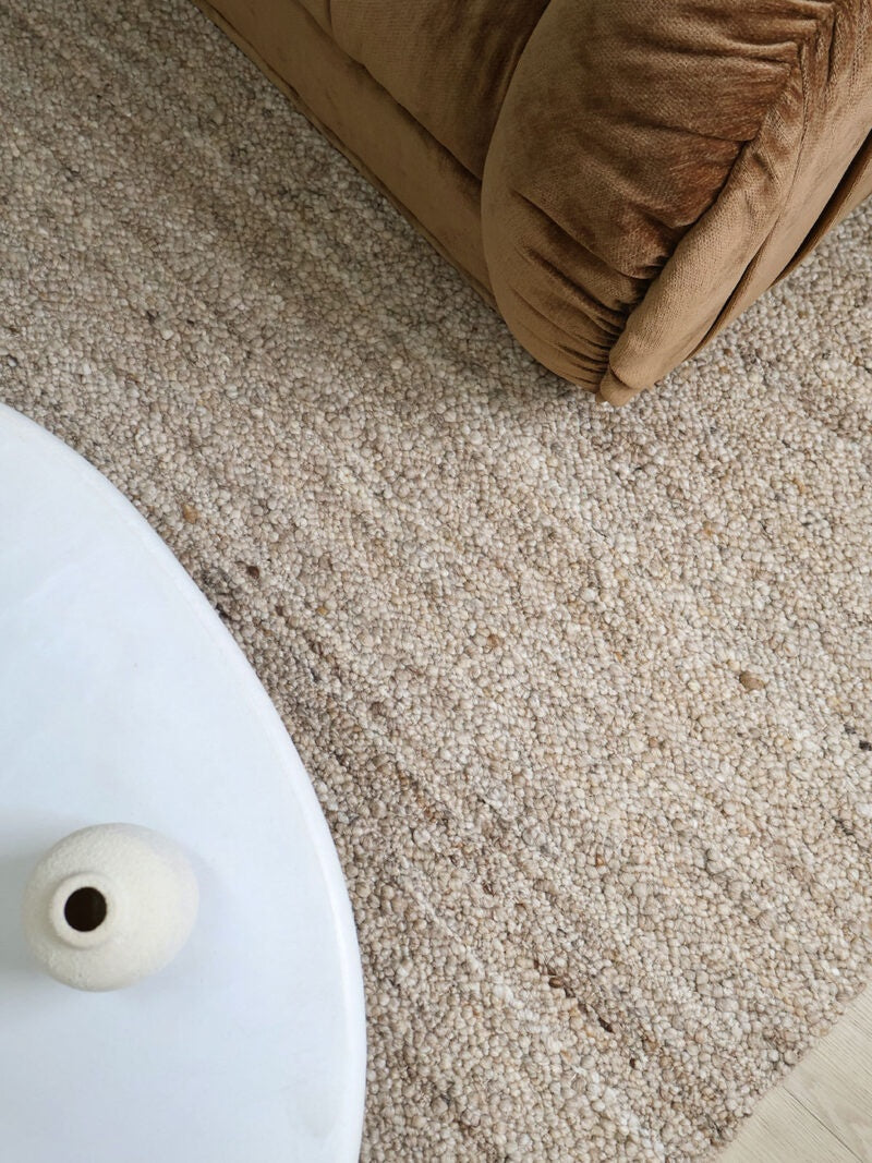 A close-up, high-angle view of a textured beige area rug in a living room setting, with the corner of a brown velvet sofa and a white side table visible.