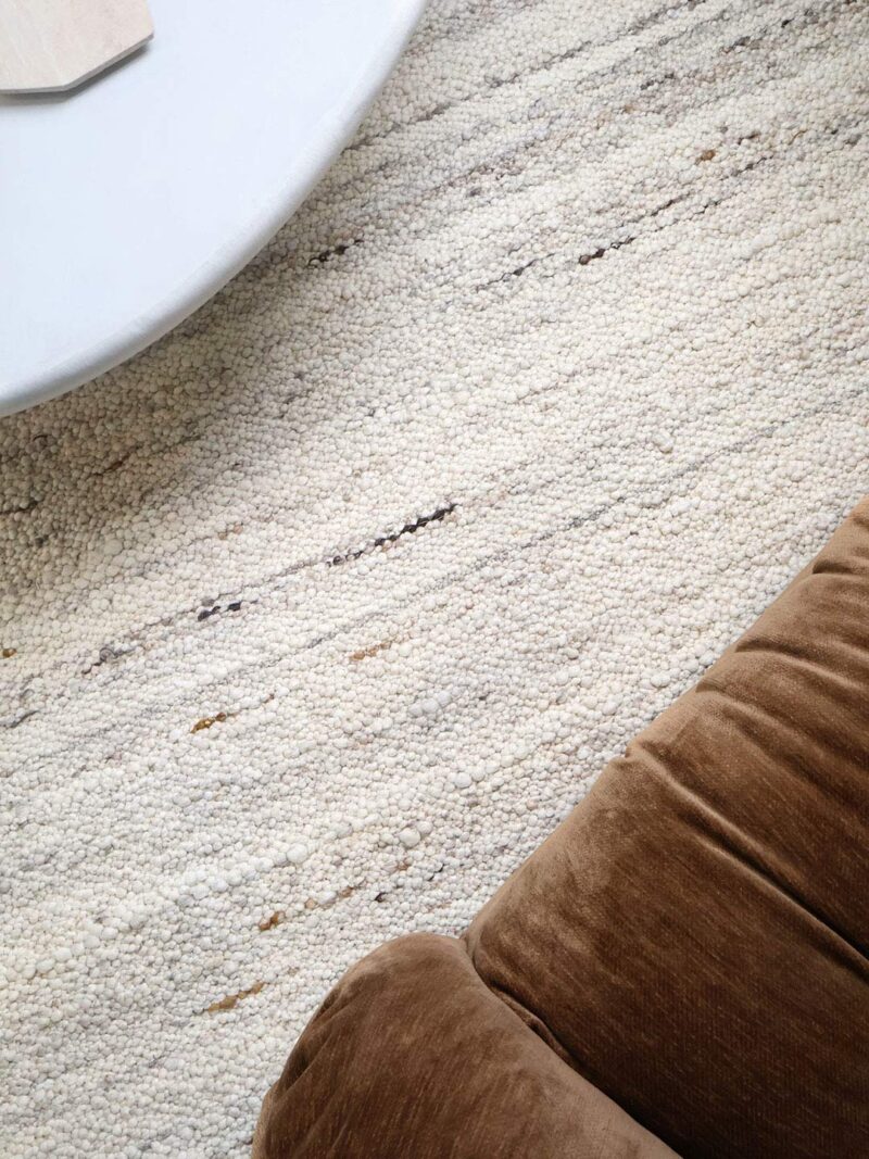A close-up of a textured cream and beige area rug with subtle horizontal woven stripes, styled in a modern interior with a brown velvet chair and a white table edge visible.